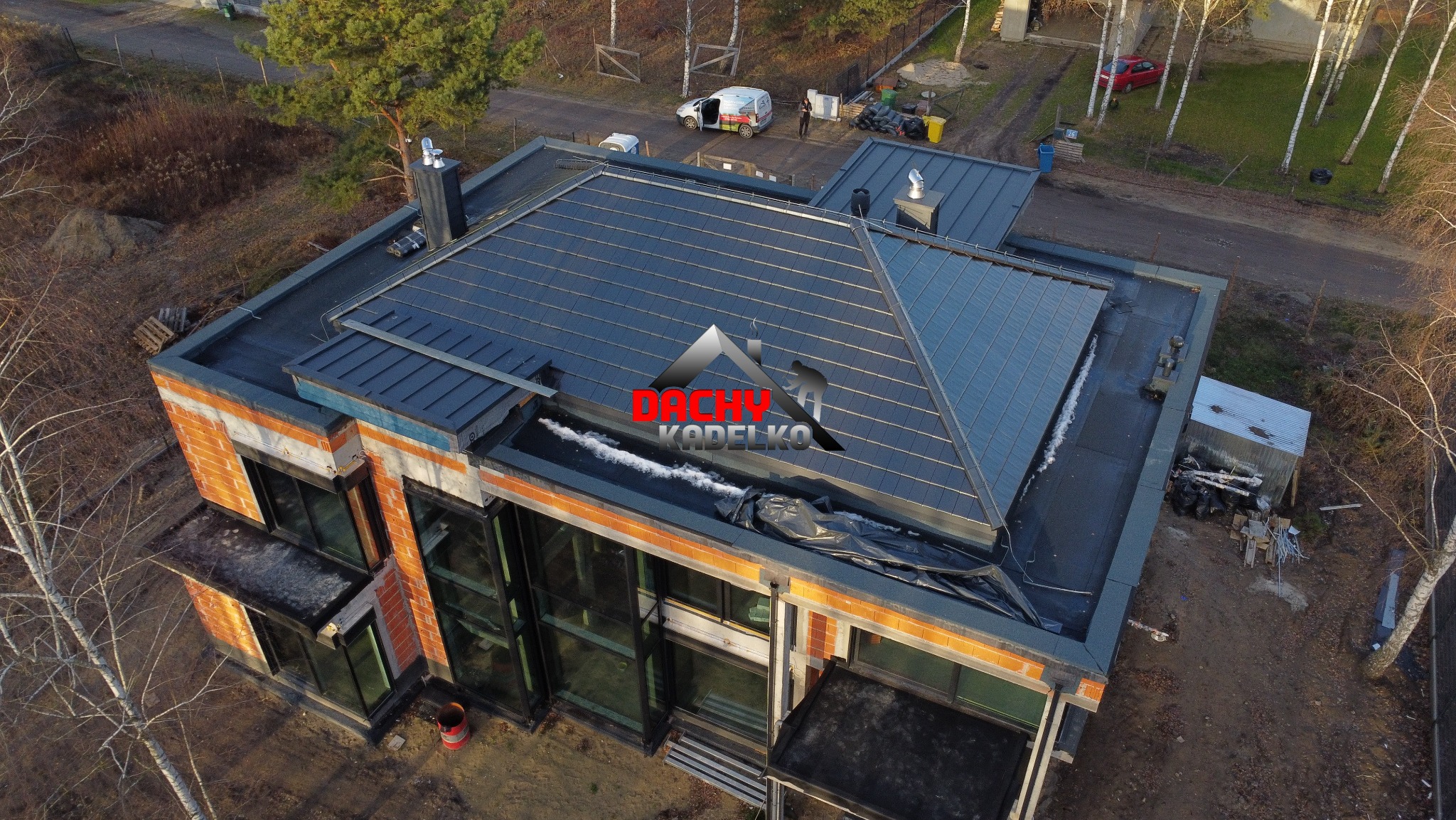 Widok z góry na nowoczesny dom z ciemnoszarą dachówką, częściowo pokrytą śniegiem. Widoczne detale architektoniczne i otoczenie budynku. Ujęcie z drona.