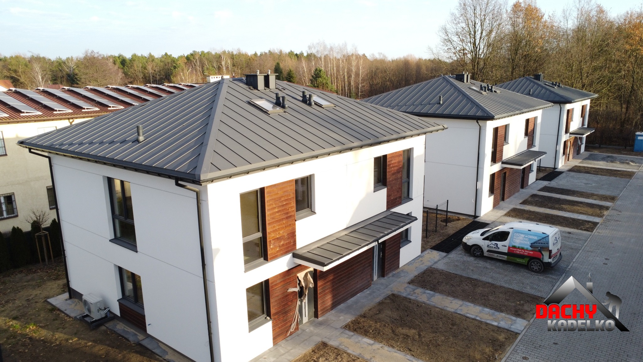 Nowoczesne domy jednorodzinne z szarym dachem z blachy na rąbek stojący, widok z góry. Na dachu okna połaciowe i kominy. Na podjeździe zaparkowany samochód firmy dekarskiej.