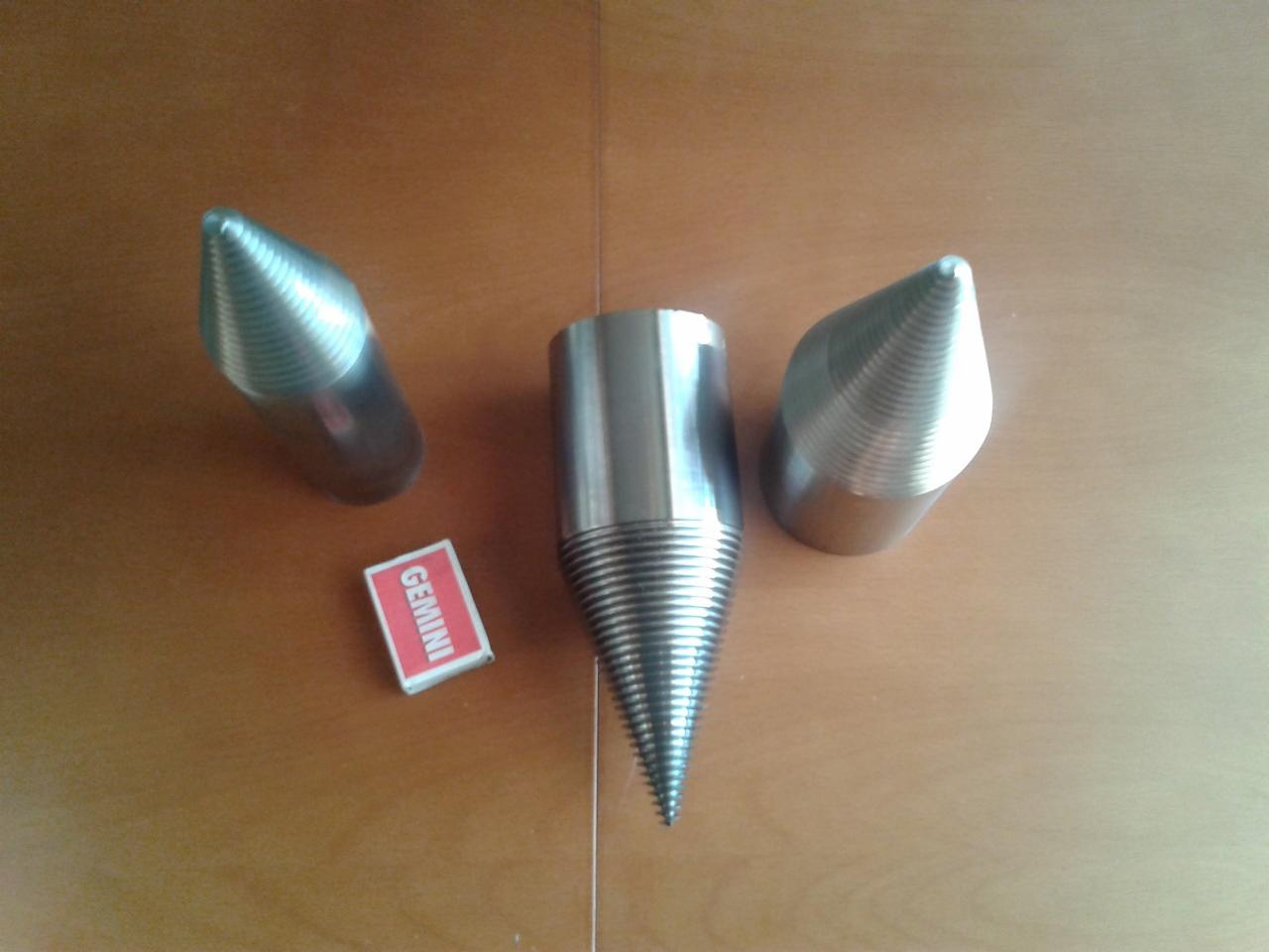 Trzy metalowe stożki z gwintem, prawdopodobnie wiertła do drewna, leżące na drewnianym blacie obok pudełka zapałek Gemini dla skali.