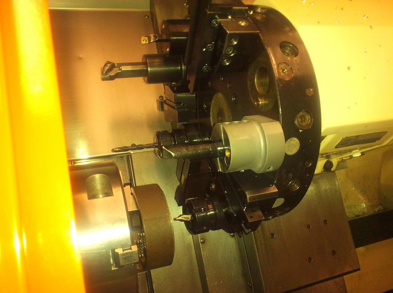 Wnętrze obrabiarki CNC z widocznymi narzędziami skrawającymi, wiertłem i uchwytami mocującymi, oświetlonym żółtym światłem.