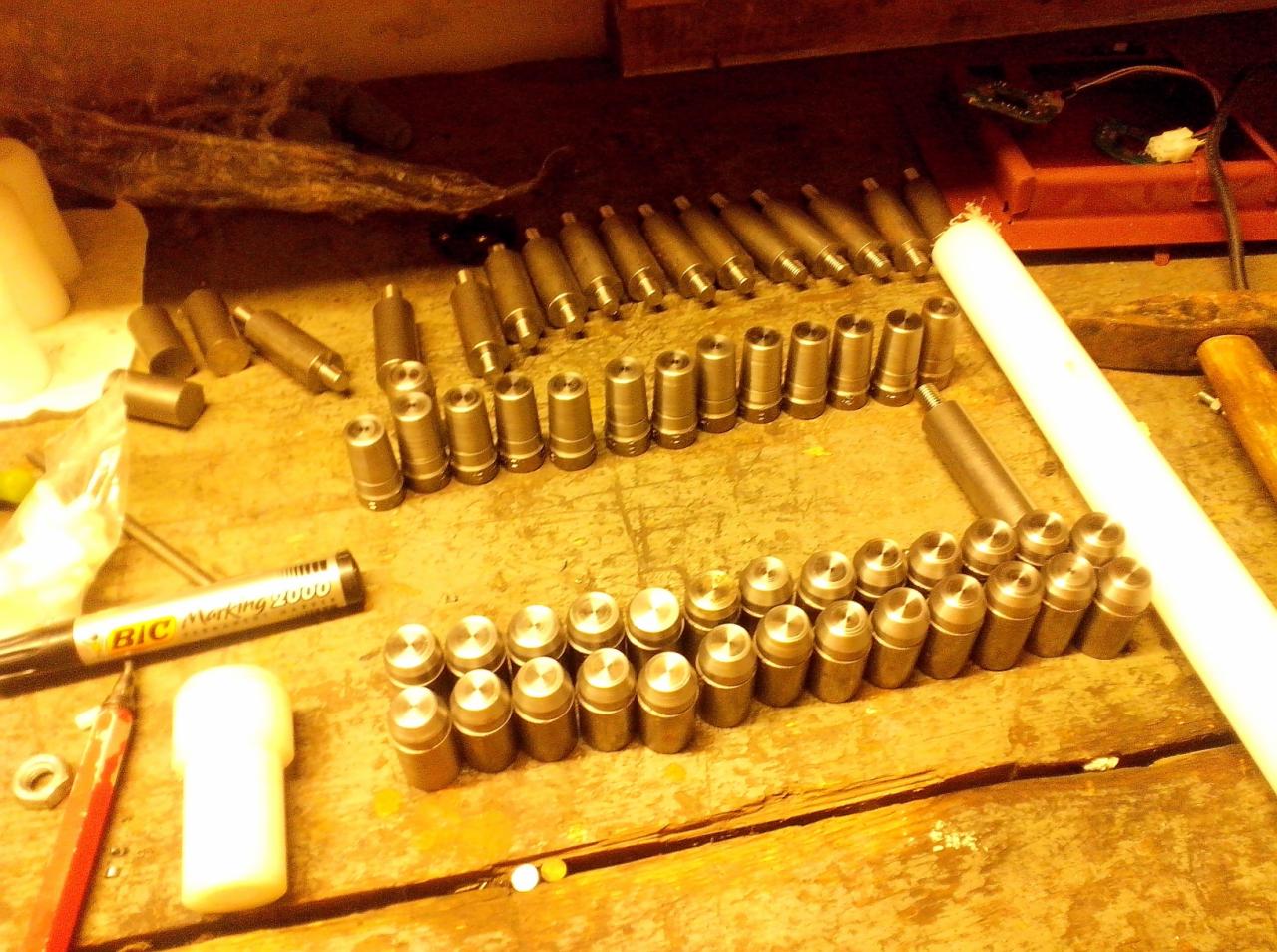 Rzędy metalowych elementów z okrągłymi główkami ułożone na drewnianym stole warsztatowym, obok leżą narzędzia i marker.