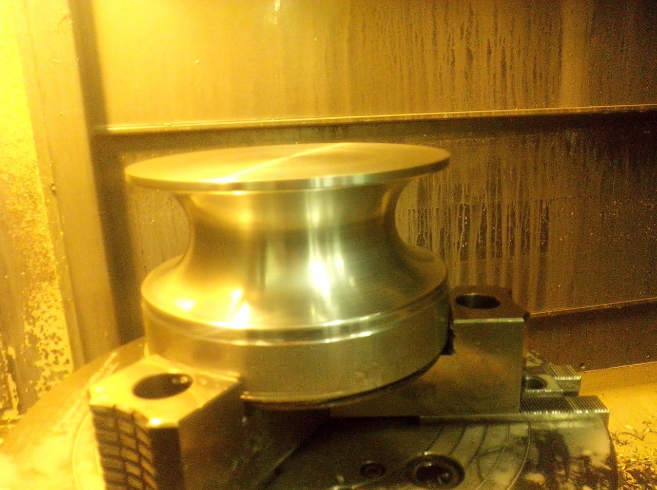 Metalowy element w kształcie szpuli, obrobiony na tokarce, zamocowany w uchwycie, widoczne ślady chłodziwa na ścianach obrabiarki.