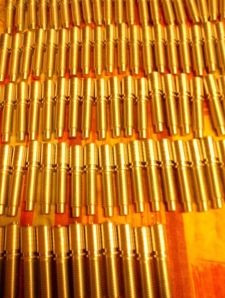 Partia złotych, gwintowanych metalowych elementów ułożonych rzędami na pomarańczowym tle, widoczne ślady obróbki.