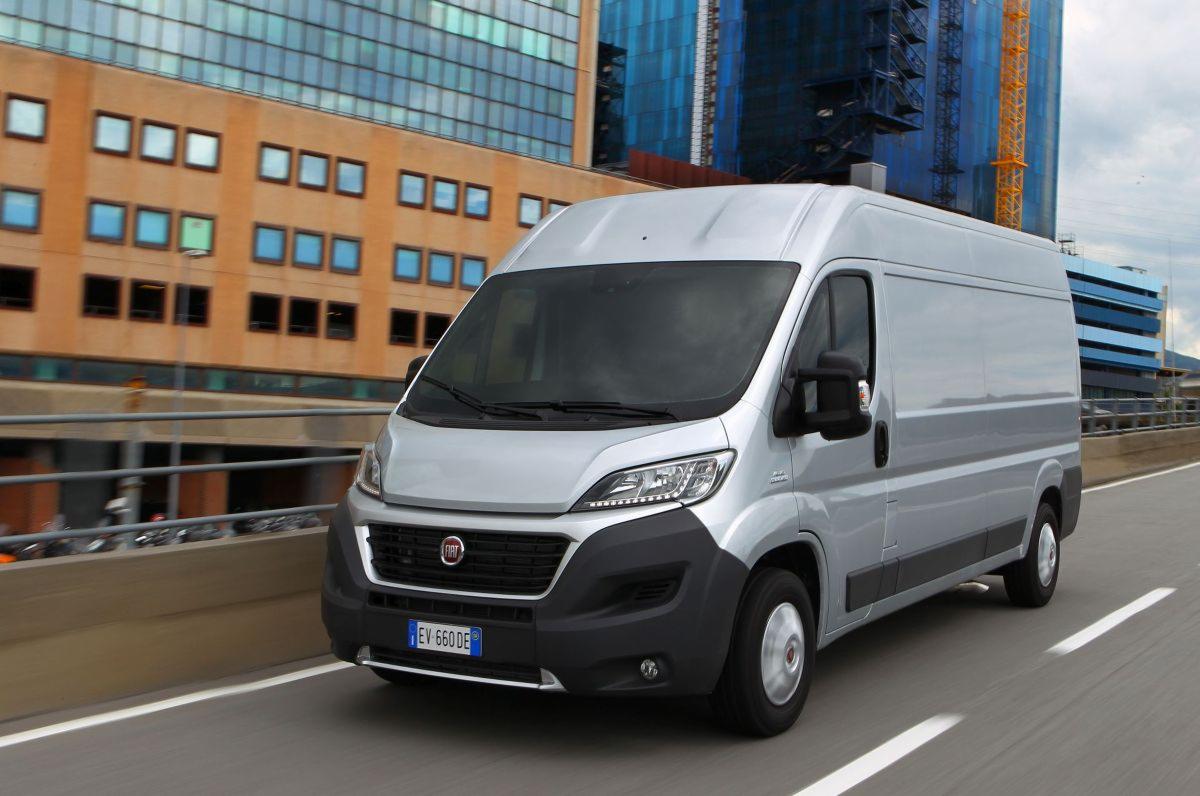 Srebrny van Fiat Ducato w ruchu na tle nowoczesnej architektury miejskiej, w oddali widoczny budynek w budowie z żółtym dźwigiem.