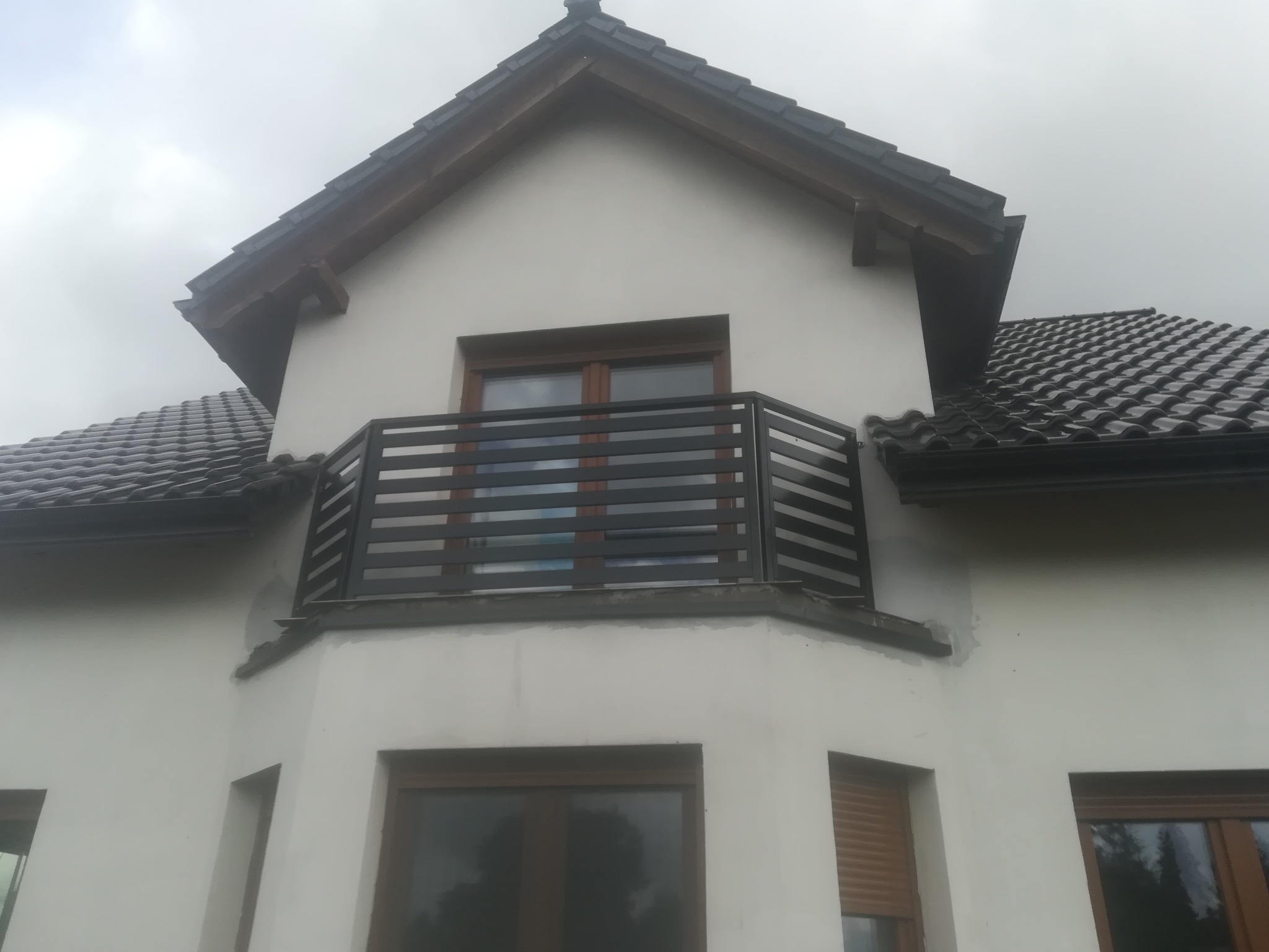 Elewacja domu z balkonem zabezpieczonym poziomą, metalową barierką w kolorze grafitowym, widoczne okna z drewnianą ramą i dach pokryty ciemną dachówką.