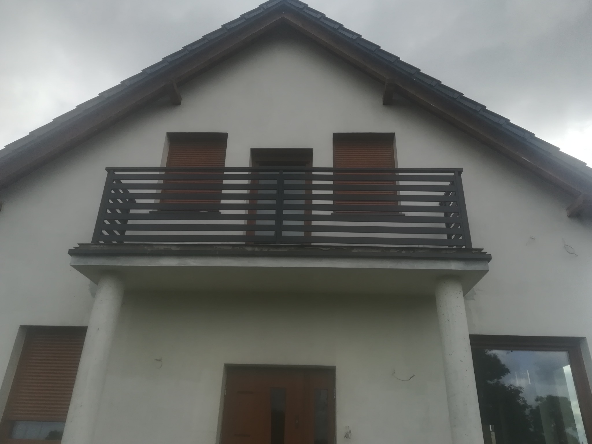 Balkon z poziomą, ciemną balustradą na tle białej fasady domu z brązowymi roletami i ciemną dachówką