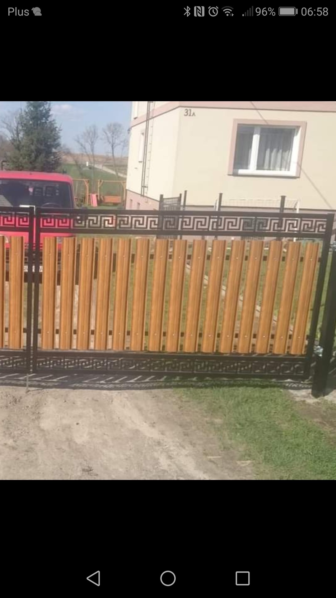 Nowoczesna brama posesyjna z metalową ramą i drewnopodobnymi pionowymi panelami, ozdobiona greckim wzorem meandrycznym, na tle budynku z numerem 31A i fragmentem czerwonego samochodu.