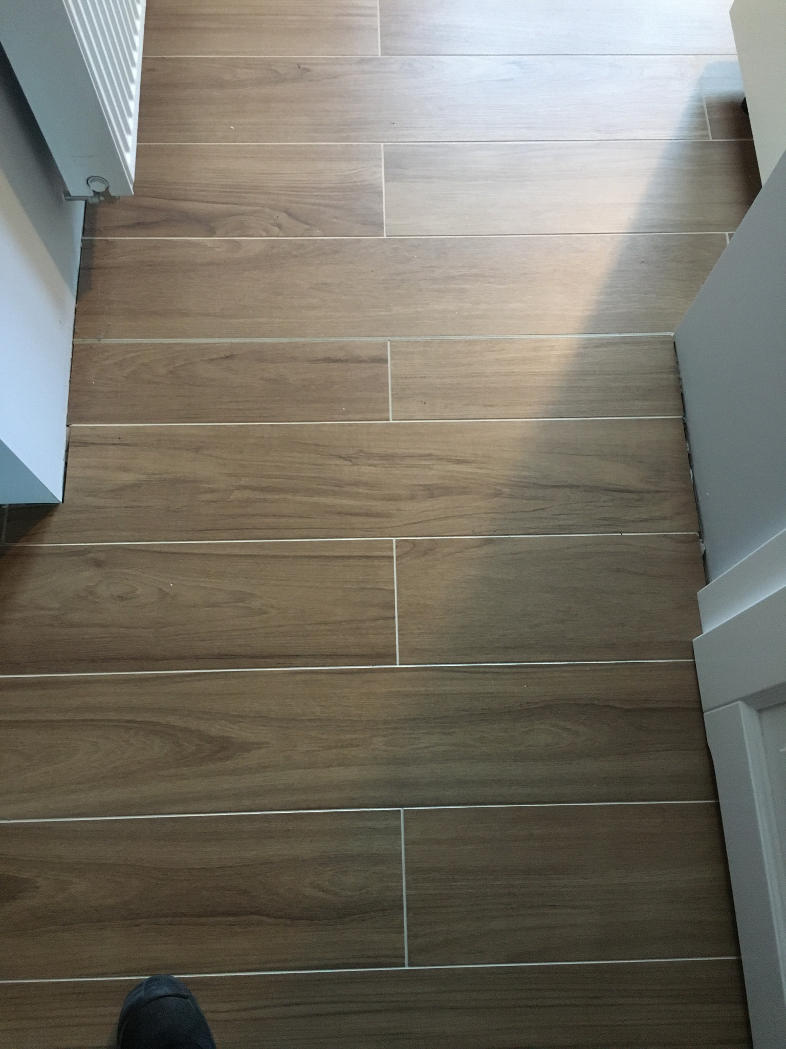 Ułożona podłoga z brązowych płytek imitujących drewno, widoczny fragment grzejnika i buta w dolnej części kadru.