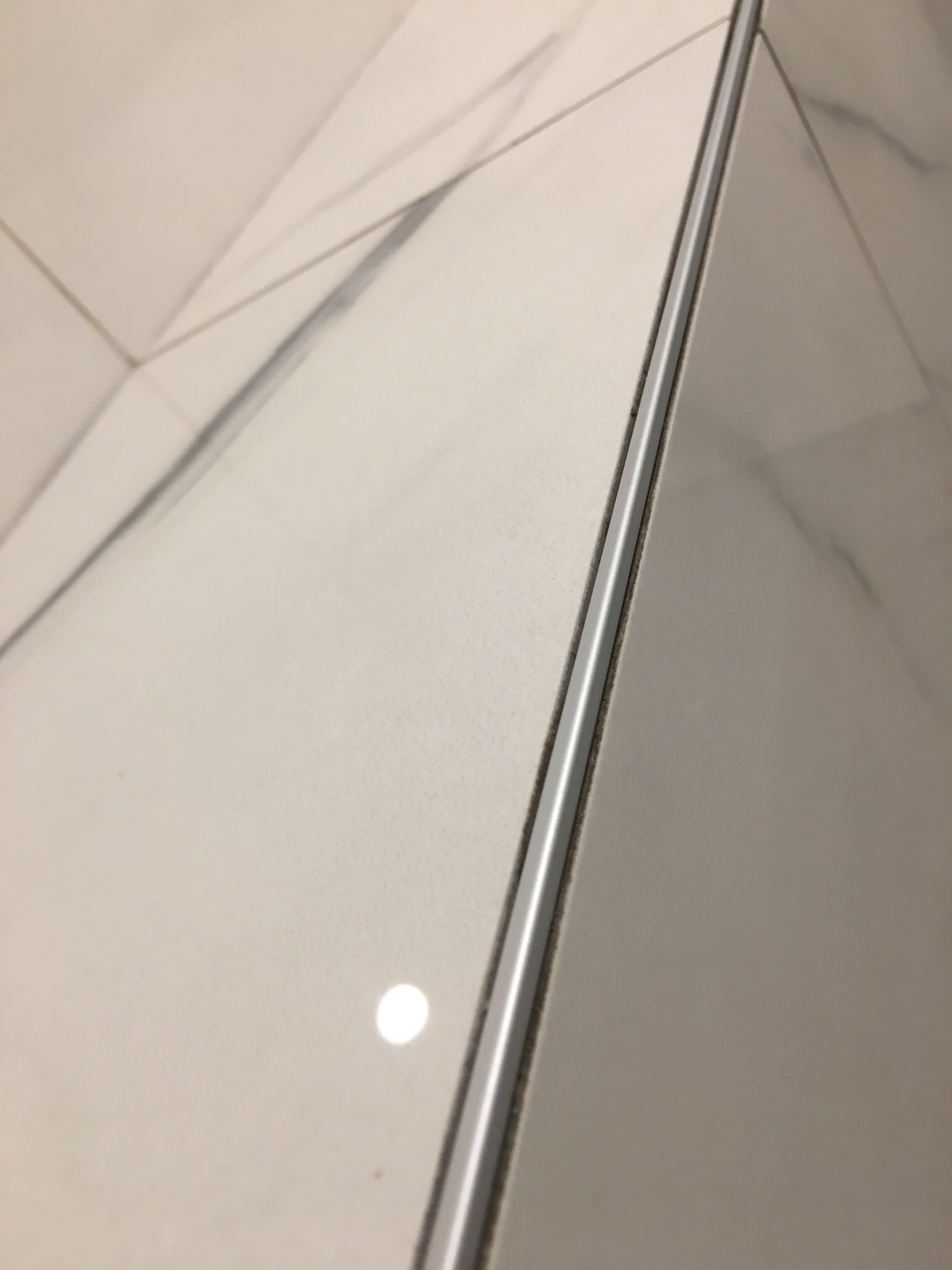 Położone jasne płytki imitujące marmur z szarą fugą i srebrną listwą wykończeniową.