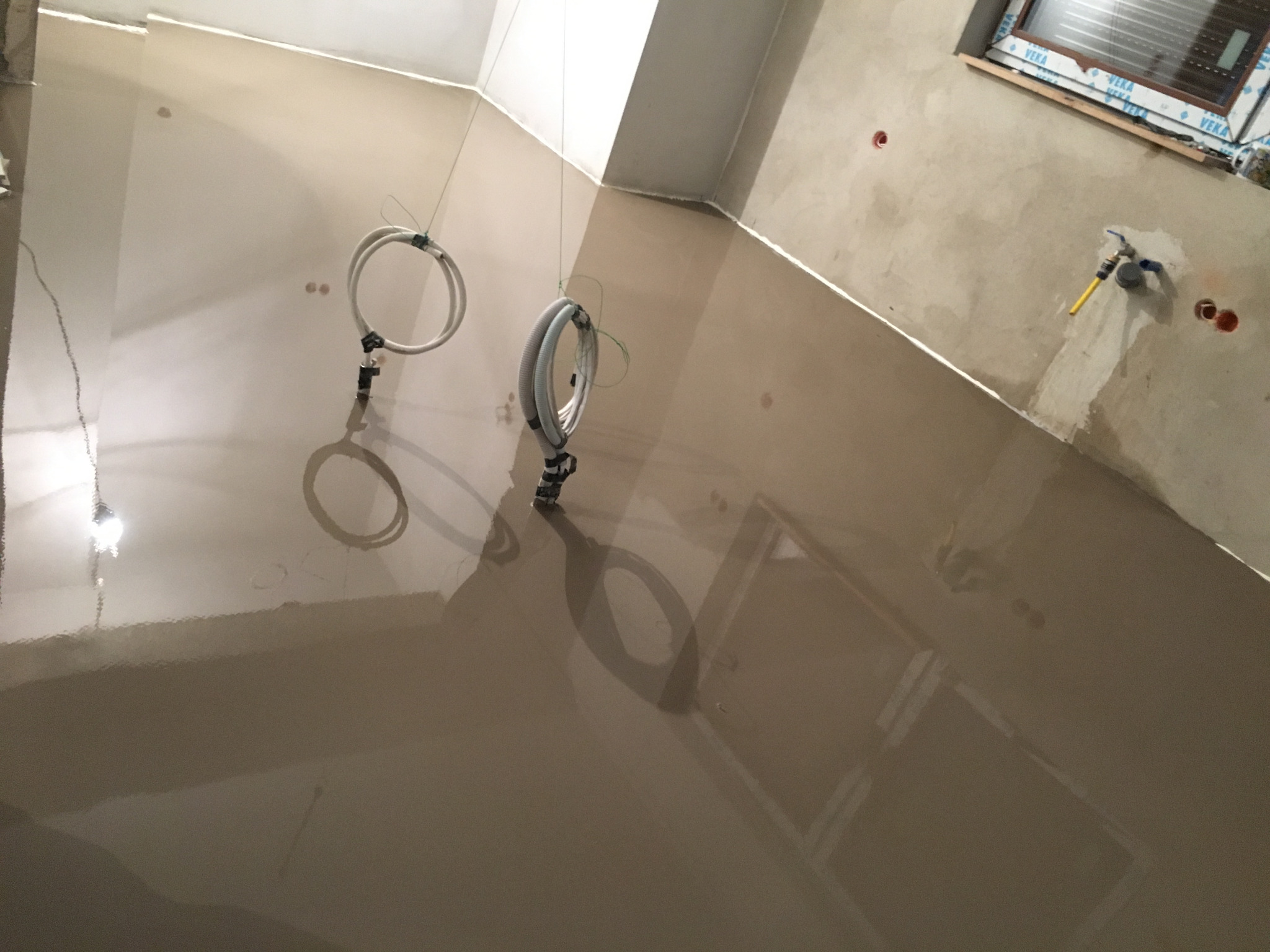 Wylewka samopoziomująca w łazience w trakcie prac, widoczne wystające rury instalacji wodnej i okno w tle.