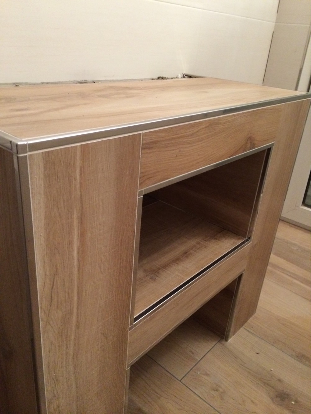 Niedokończona zabudowa umywalki z drewnopodobnych płytek ceramicznych z aluminiowymi profilami, widoczne niedociągnięcia przy łączeniu ze ścianą.