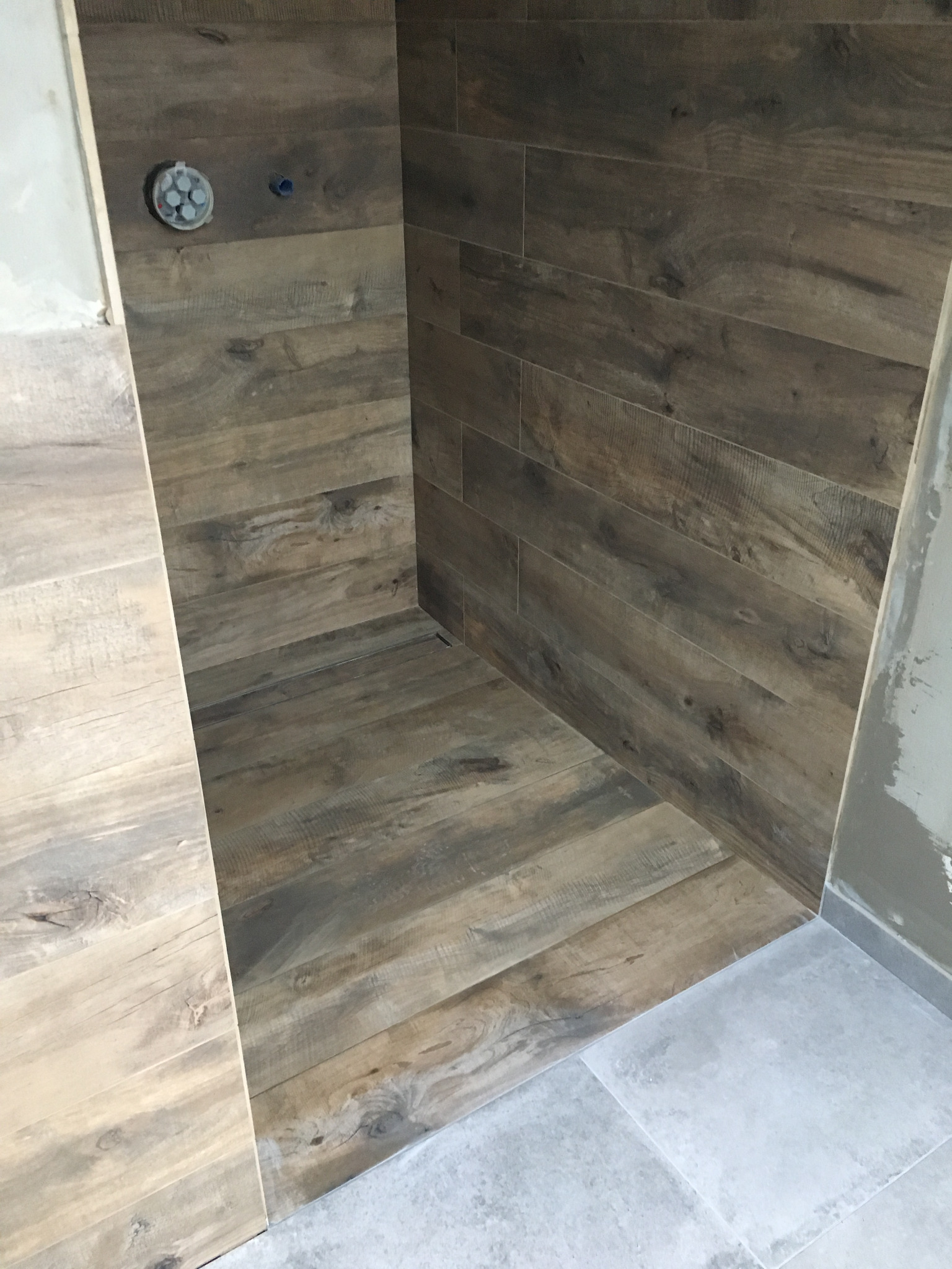 Wnęka prysznicowa wyłożona płytkami imitującymi drewno; widoczne przyłącza wodne i fragment szarej podłogi poza kabiną.
