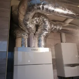 Dwa białe piece gazowe marki Vaillant zamontowane na ścianie z imitacji drewna, połączone srebrnymi, zaizolowanymi przewodami wentylacyjnymi.