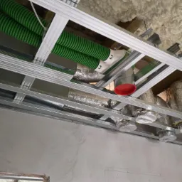 Widok na sufit podwieszany w trakcie montażu instalacji wentylacyjnej z zielonymi rurami karbowanymi i srebrnymi kanałami izolowanymi folią aluminiową, widoczna konstrukcja z profili metalowych.