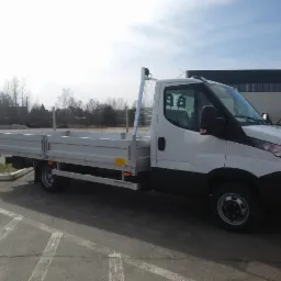 Biały samochód dostawczy z otwartą skrzynią ładunkową, zaparkowany na parkingu z widocznymi liniami, w tle budynek z ciemnymi oknami i częściowo widoczny inny pojazd.