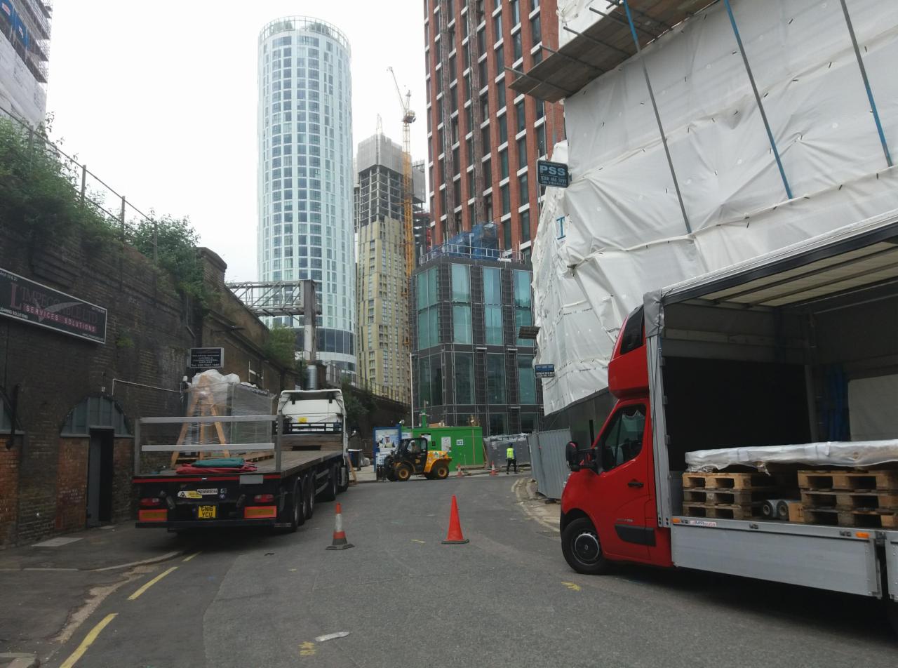 Ciężarówki dostarczają materiały budowlane na plac budowy w miejskiej scenerii z wysokimi budynkami w tle.