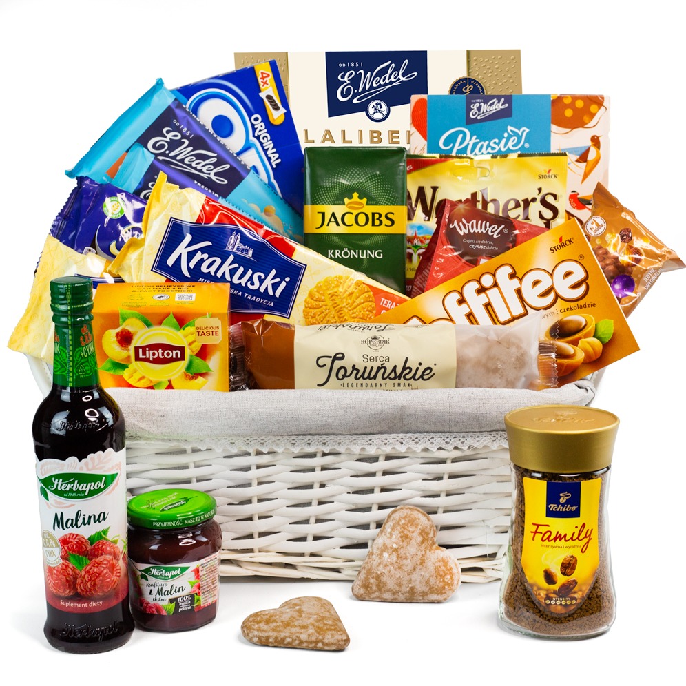 Biały wiklinowy kosz wypełniony różnorodnymi słodyczami, kawą, herbatą i syropem owocowym, z piernikami w kształcie serca na pierwszym planie.