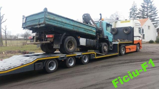 Ciężarówka wywrotka załadowana na niskopodwoziową lawetę transportową, z ciągnikiem siodłowym z tyłu, na placu manewrowym.