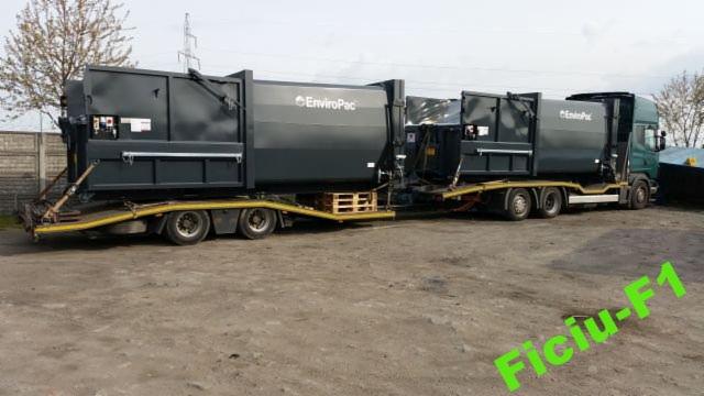Dwa zestawy transportowe z czarnymi kontenerami EnviroPac na niskopodwoziowych przyczepach, jeden kontener zabezpieczony paletą, widok z boku na szarym placu.
