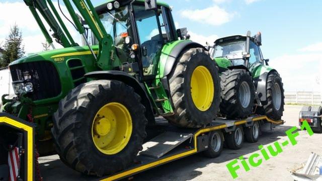 Dwa zielone traktory John Deere na niskopodwoziowej przyczepie z najazdami, widoczne z boku, w tle błękitne niebo.