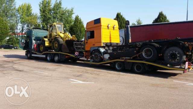 Żółta ładowarka na platformie niskopodwoziowej ciągniętej przez pomarańczowy ciągnik siodłowy, widok boczny na parkingu.