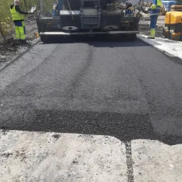 Maszyna rozkładająca asfalt na drodze, pracownicy w odblaskowych ubraniach nadzorują proces, widoczne świeżo rozłożone warstwy asfaltu i fragmenty starej nawierzchni.