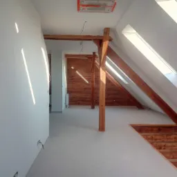 Poddasze po remoncie z drewnianą konstrukcją więźby dachowej, świeżo pomalowane ściany w kolorze białym, widoczne kable elektryczne i fragment muru z czerwonej cegły w tle.