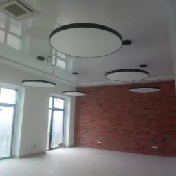 Wnętrze pomieszczenia z sufitami napinanymi, oświetleniem w postaci okrągłych lamp, ścianą z cegły i dużymi oknami zabezpieczonymi folią ochronną.