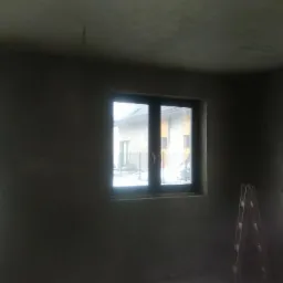 Pokój w stanie surowym po tynkowaniu, widoczne trzy okna z widokiem na zewnątrz, drabina i wiadro na stołku, ściany szare, nierówne.