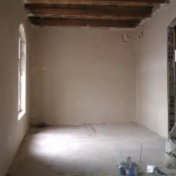 Wnętrze pokoju w trakcie prac wykończeniowych: świeżo nałożony tynk na ścianach, widoczne drewniane belki stropowe, okno z łukowym nadprożem, sprzęt tynkarski na podłodze.