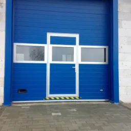 Niebieska segmentowa brama z wbudowanymi oknami i drzwiami wejściowymi, wykończona taśmą ostrzegawczą, zamontowana w surowym budynku z bloczków betonowych.