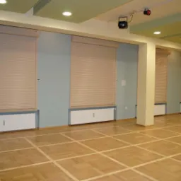 Wnętrze sali z parkietem w kratkę i beżowymi roletami zewnętrznymi częściowo zasłoniętymi, widoczne grzejniki pod oknami, oświetlenie sufitowe i kula dyskotekowa.