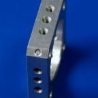 Detal z obrobionego aluminium z rzędem otworów na krawędzi, widoczne ślady obróbki skrawaniem, niebieskie tło.