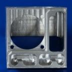 Precyzyjnie obrobiony metalowy element z widocznymi frezowanymi wgłębieniami o różnych kształtach, w tym okrągłym i prostokątnym, na jednolitym niebieskim tle.