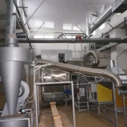 Wnętrze hali produkcyjnej z zainstalowaną przemysłową instalacją wentylacyjną i odciągową z metalowymi rurami, filtrem cyklonowym i elementami sterującymi.