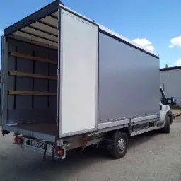 Pojazd dostawczy z otwartą przestrzenią ładunkową, widok z tyłu, z elementami mocującymi wewnątrz i częściowo zasuniętą plandeką z boku, na szarym placu.