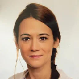 Portret kobiety z ciemnymi włosami splecionymi w warkocz, uśmiechająca się delikatnie, na jasnym tle.