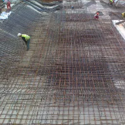 Siatka zbrojeniowa ułożona na betonowym fundamencie, pracownicy w kaskach i kamizelkach odblaskowych w trakcie montażu zbrojenia.