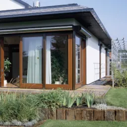 Drewniane, przesuwne drzwi tarasowe z roletami zewnętrznymi w białym domu z drewnianym tarasem i zadbanym ogrodem.