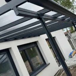 Nowoczesna pergola z szarym stelażem i przezroczystym zadaszeniem, przylegająca do białego budynku z oknami w szarych ramach. Widoczna drabina i elementy instalacji.