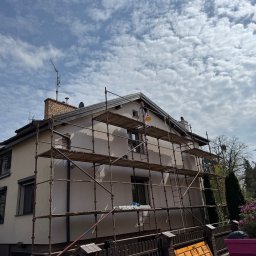 Remokompleks Michał Motyl - Malowanie elewacji domu z rusztowania, pracownik z żółtym wiadrem na wysokości. Widok na budynek z zewnątrz, błękitne niebo z chmurami w tle.