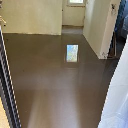 Remokompleks Michał Motyl - Wylana, mokra wylewka samopoziomująca w pomieszczeniu z oknem. Widoczne ślady po zabrudzeniach na ścianach. Część okna odbija się w mokrej powierzchni.