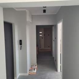 Remokompleks Michał Motyl - Szary korytarz po remoncie z drzwiami, oświetleniem sufitowym i parą butów na wycieraczce. Świeżo pomalowane ściany i minimalistyczny design.