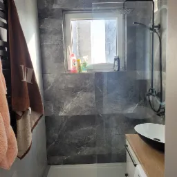 Nowoczesna łazienka z szarymi płytkami imitującymi marmur, kabiną prysznicową typu walk-in i umywalką nablatową. Okno z widokiem na budynek.