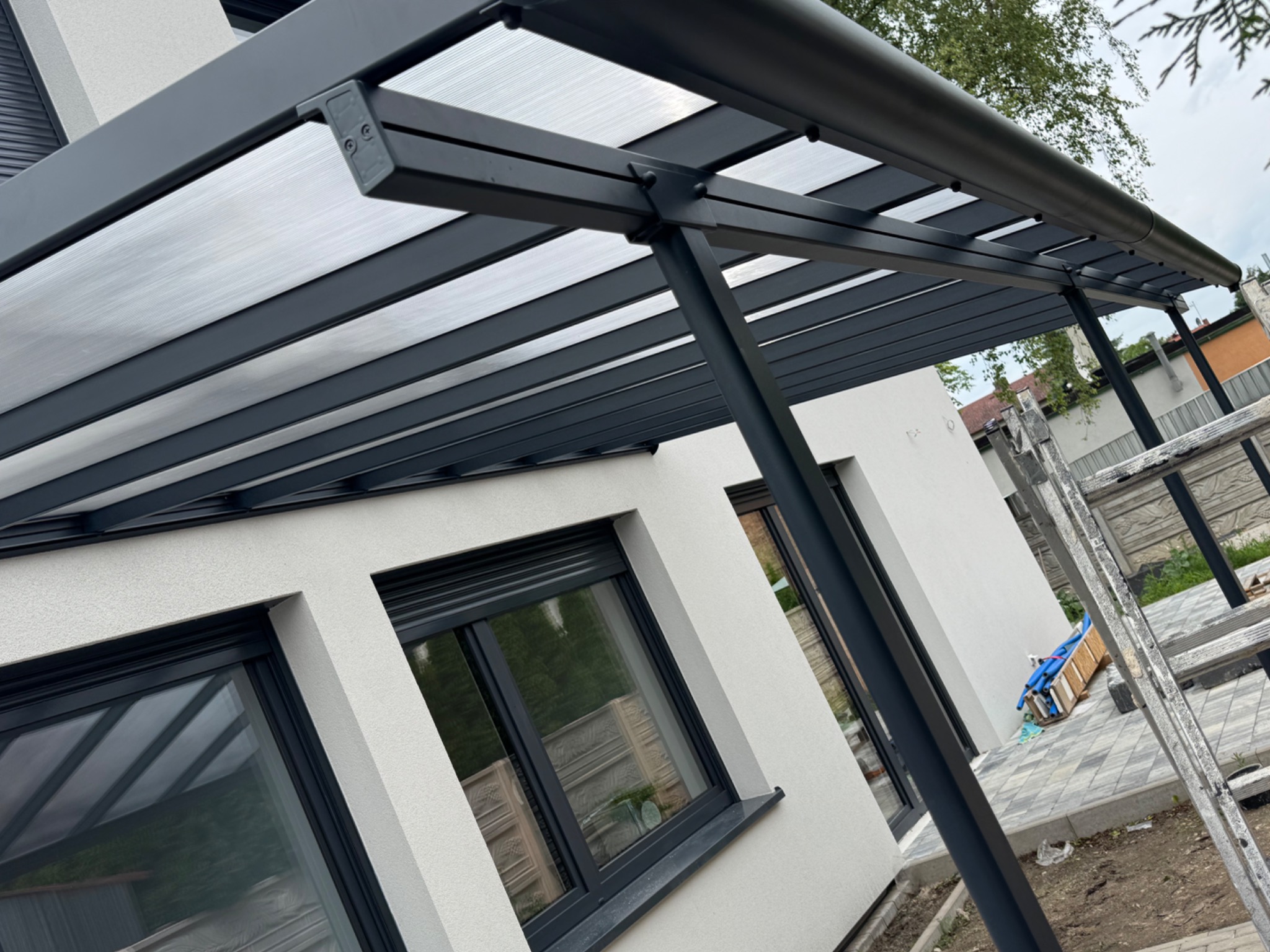 Nowoczesna pergola z szarym stelażem i przezroczystym zadaszeniem, przylegająca do białego budynku z oknami w szarych ramach. Widoczna drabina i elementy instalacji.