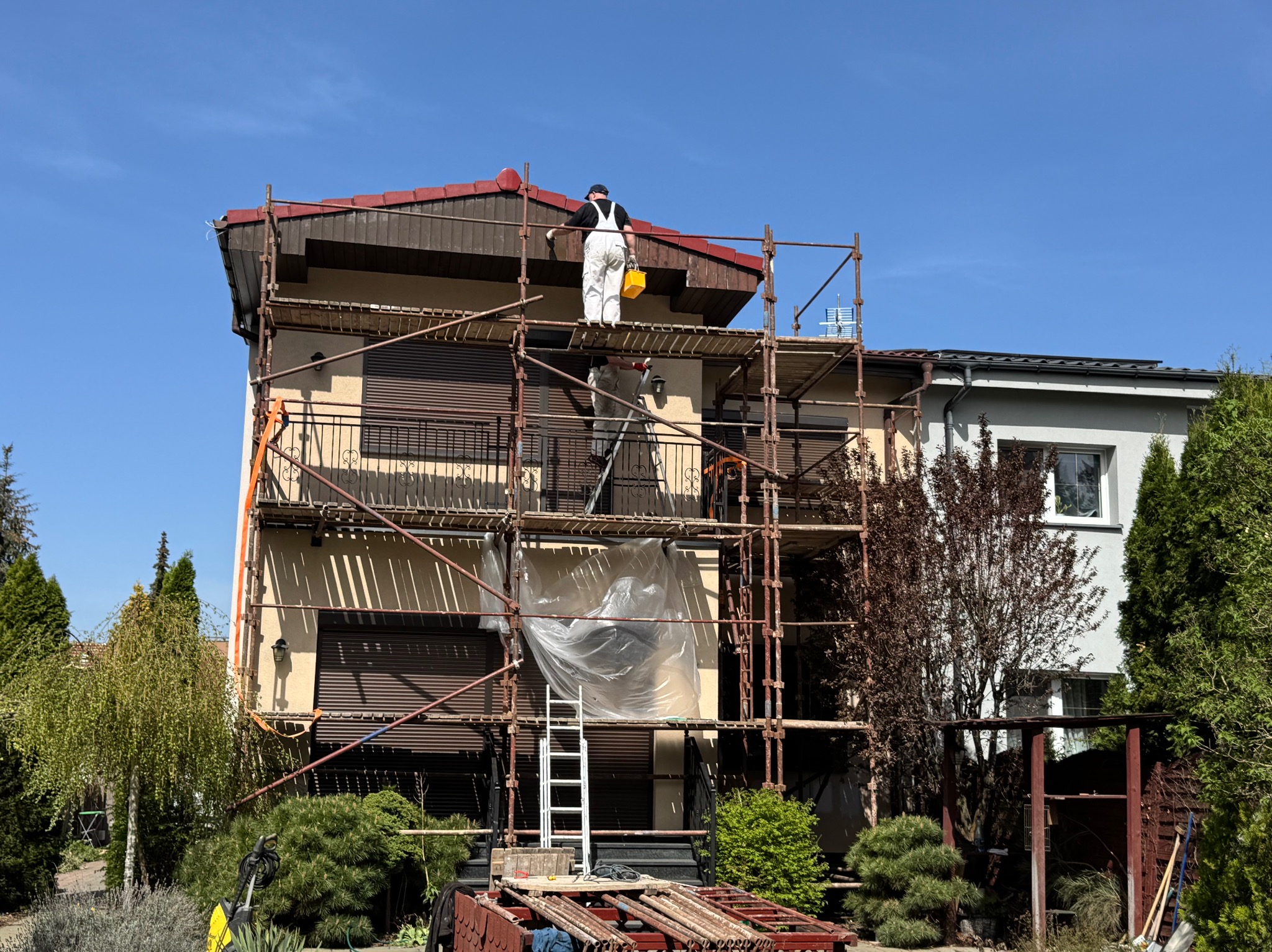 Malowanie elewacji domu jednorodzinnego z rusztowania. Pracownik w kombinezonie maluje fragment dachu. Widoczne narzędzia i zabezpieczenia.