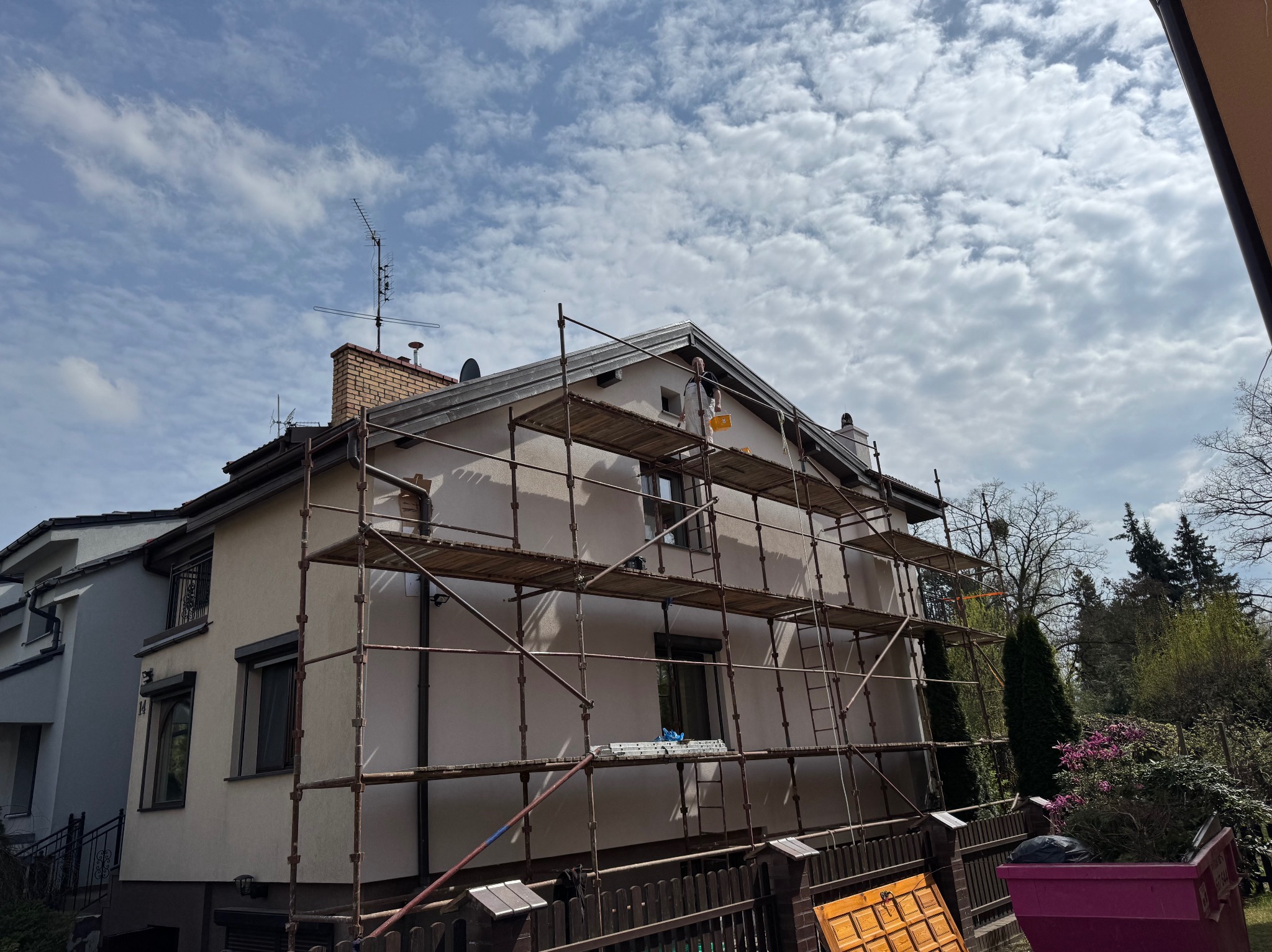 Elewacja domu jednorodzinnego w trakcie prac malarskich, widoczne rusztowania, pracownik w kombinezonie malarskim przy szczycie budynku. Błękitne niebo z obłokami.