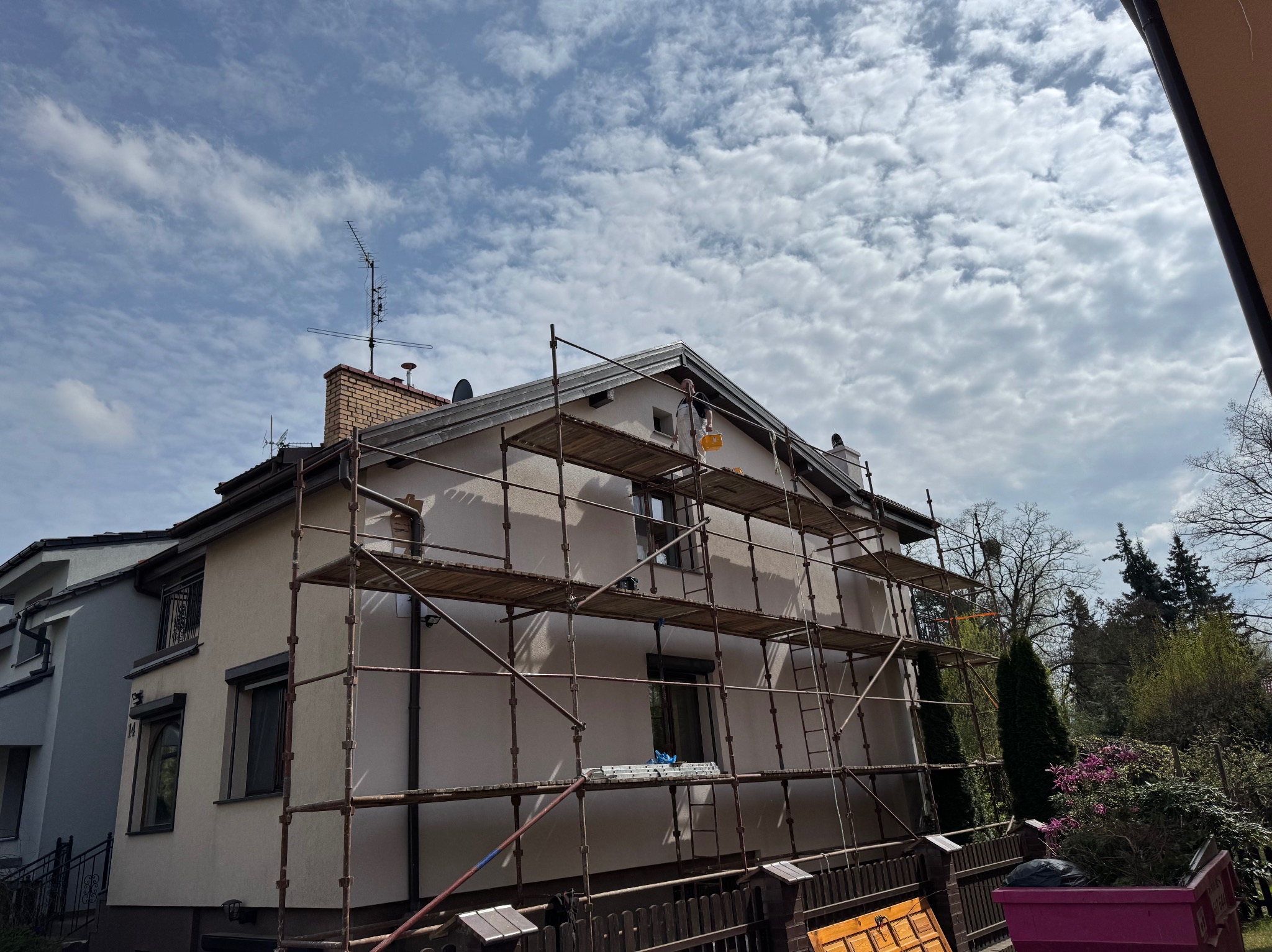 Malowanie elewacji domu z rusztowania, pracownik z żółtym wiadrem na wysokości. Widok na budynek z zewnątrz, błękitne niebo z chmurami w tle.