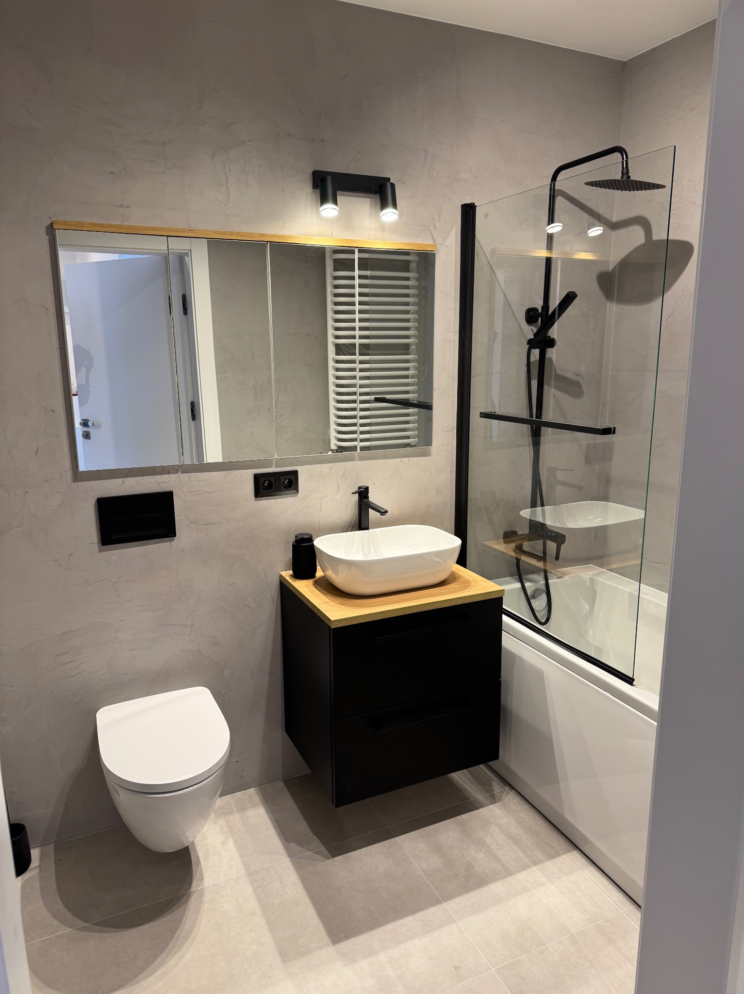 Nowoczesna łazienka w szarościach: wisząca toaleta, czarna szafka z umywalką, lustro w drewnianej ramie, kabina prysznicowa z czarną armaturą. Minimalistyczny design.