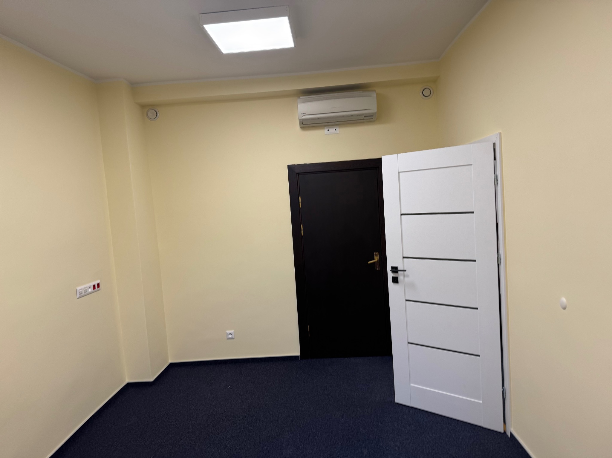 Pomalowane na jasnożółto wnętrze z ciemną podłogą, białą, nowoczesną ościeżnicą i klimatyzacją na ścianie. Widoczne gniazdka i włączniki.